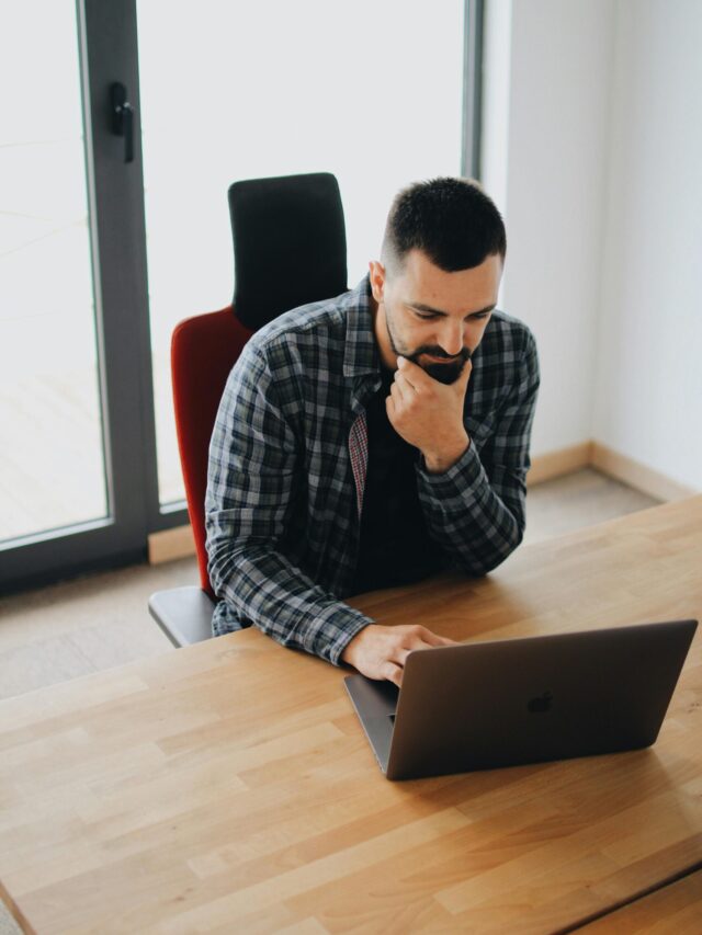 Young man with beard working on laptop at desk in a modern office setting.