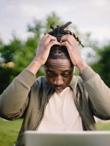 Upset young African American male freelancer clutching head with hands after failure in project while working remotely in park